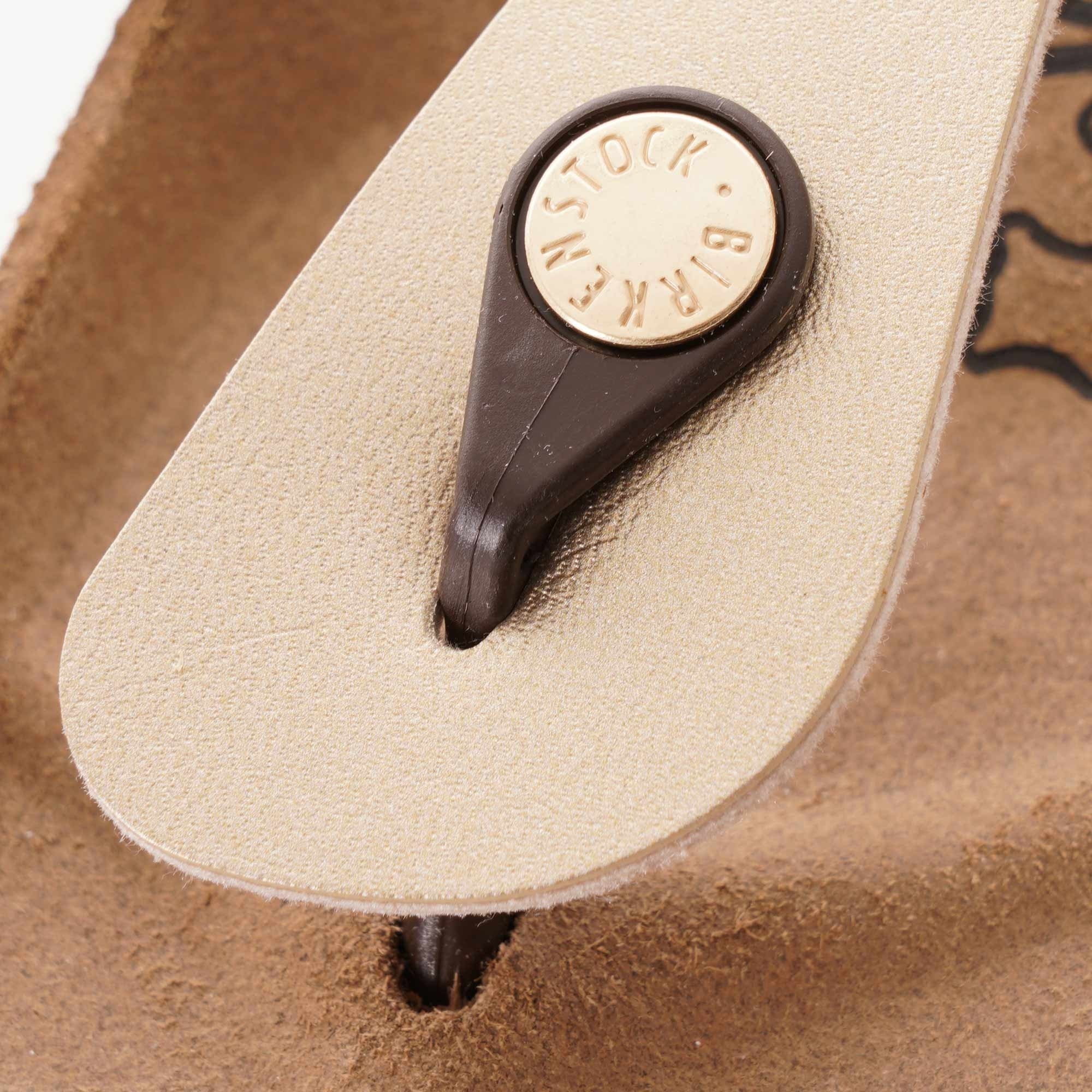 A close-up of the Birkenstock branded stud on a gold Gizeh sandal's toe post.