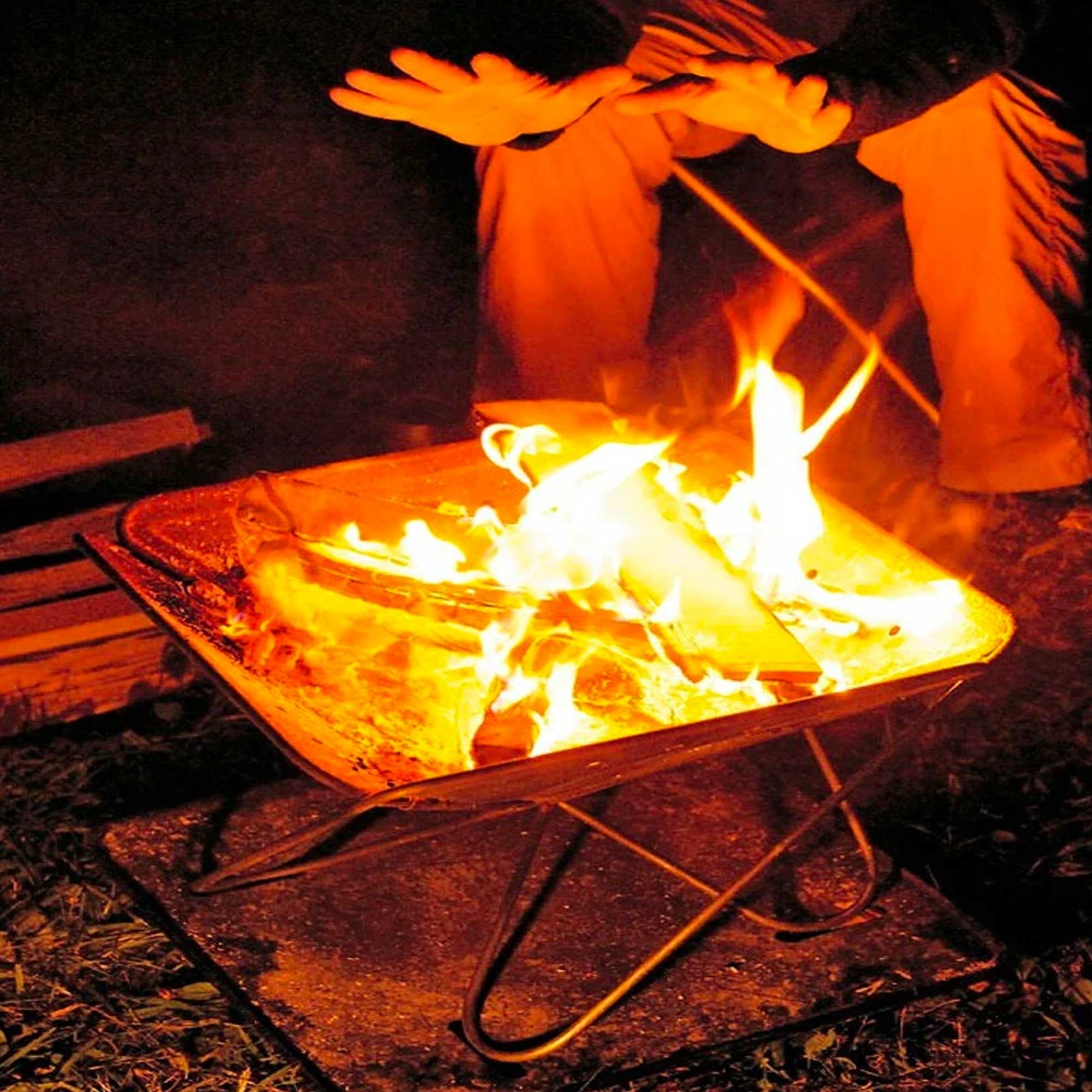 Two people warming their hands over a lit stainless steel fire pit at night.