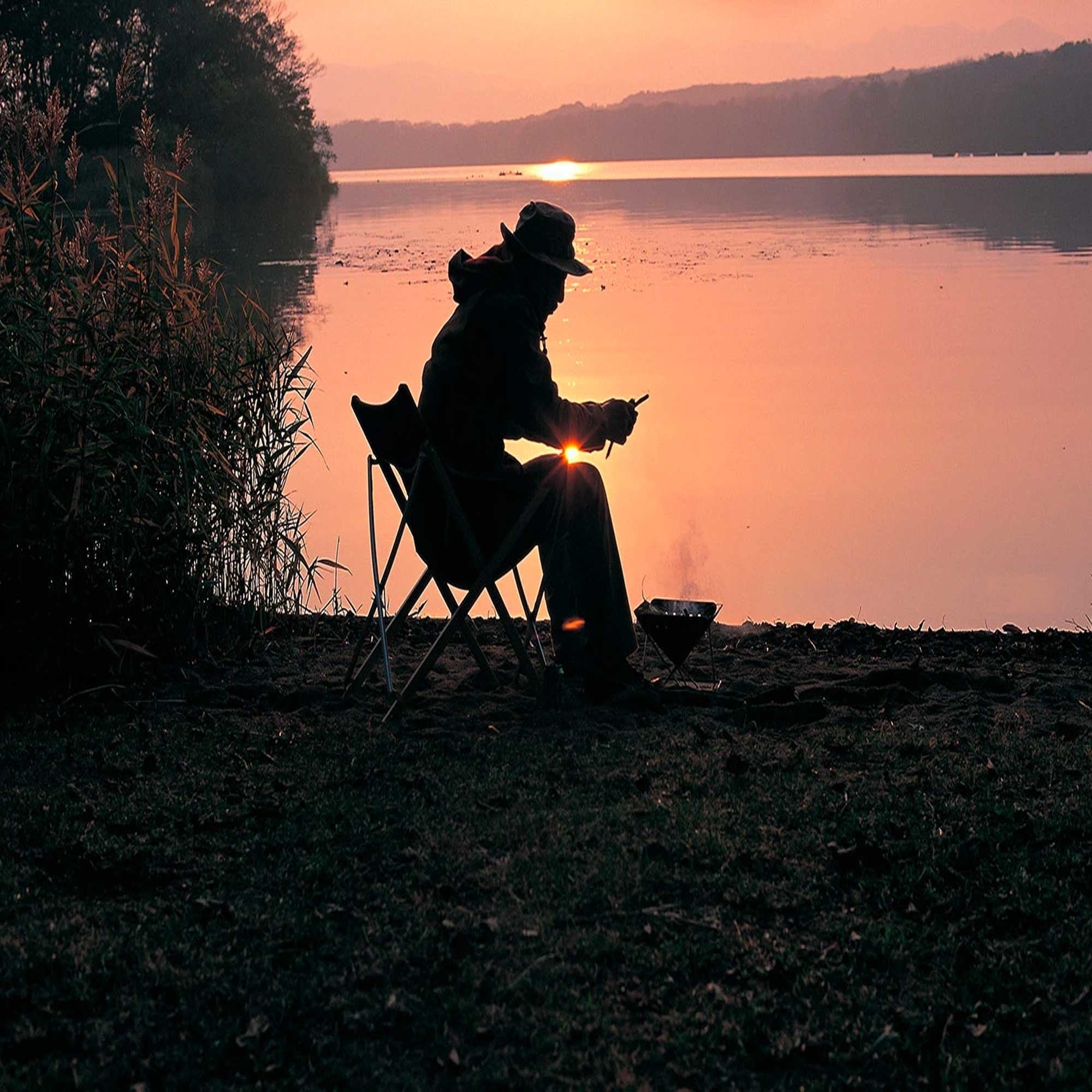 A person sitting on a chair next to a fire pit, with a lake and sunset in the background.
