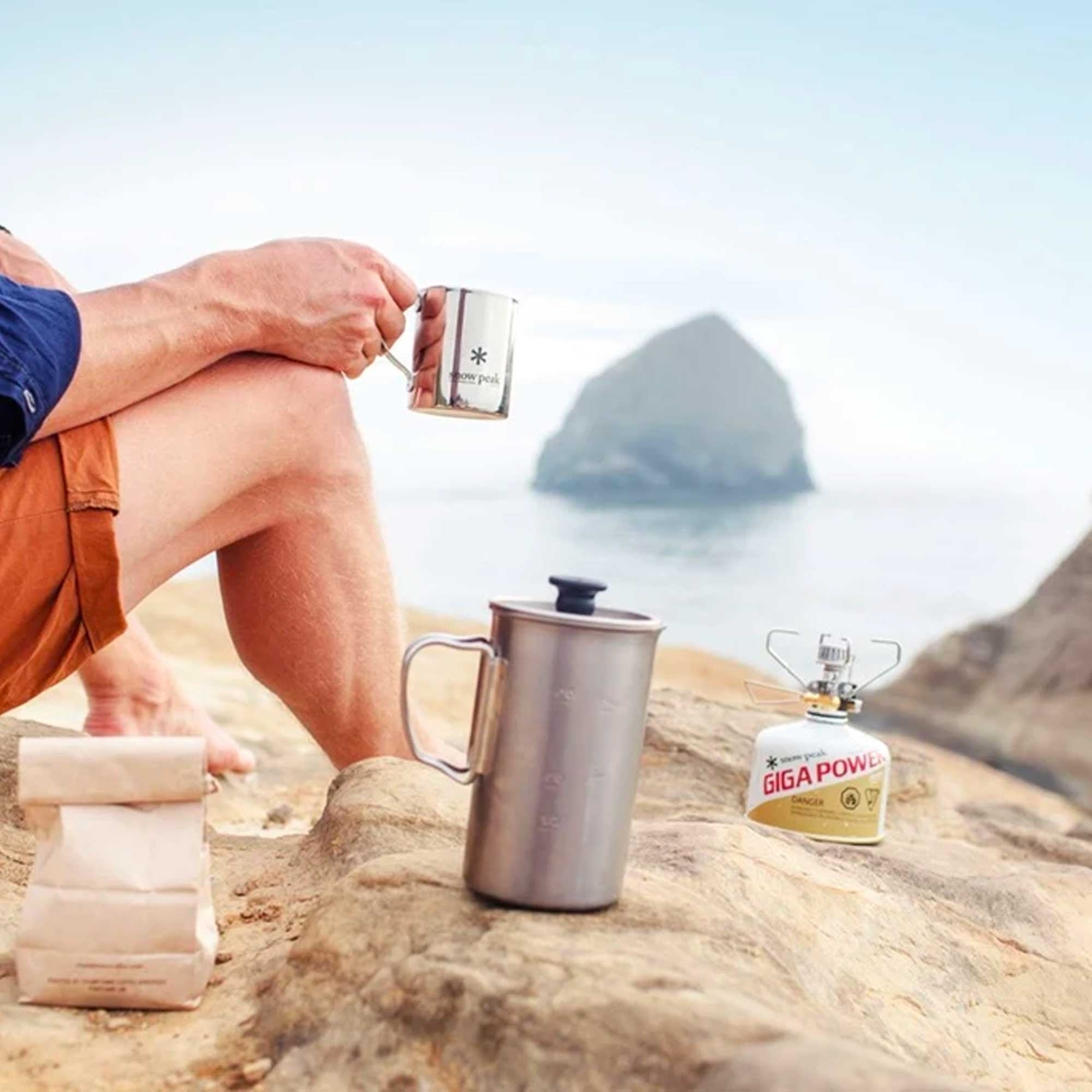 A person holds a small metal mug, with a larger coffee press and camping stove nearby.