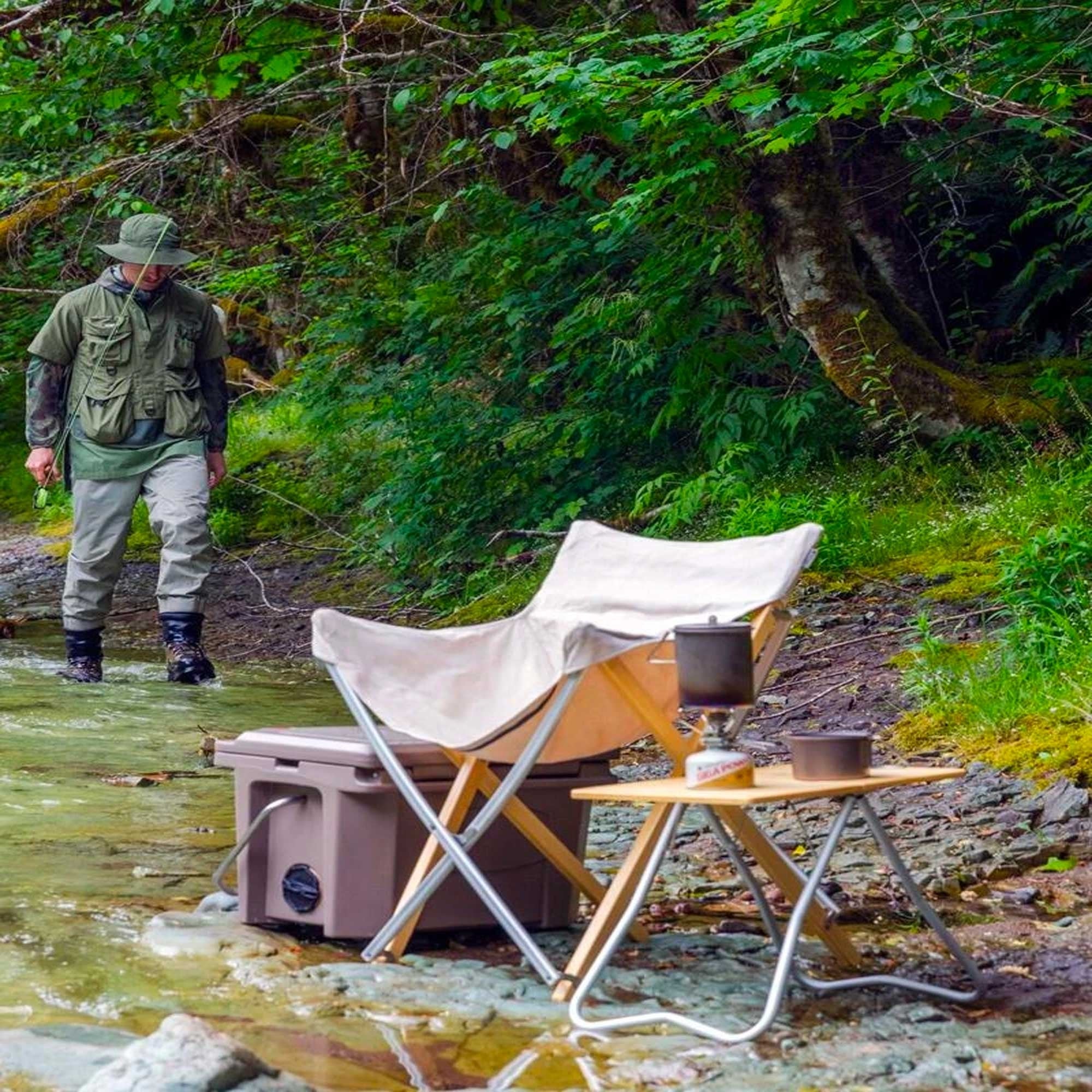 A person fly fishing in a stream, with camping gear set up on the bank.