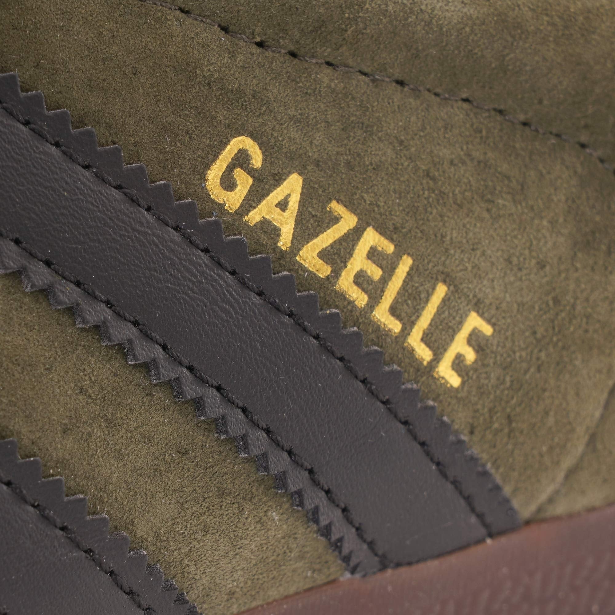 A close-up of the gold 'Gazelle' lettering on the side of an olive green trainer. 