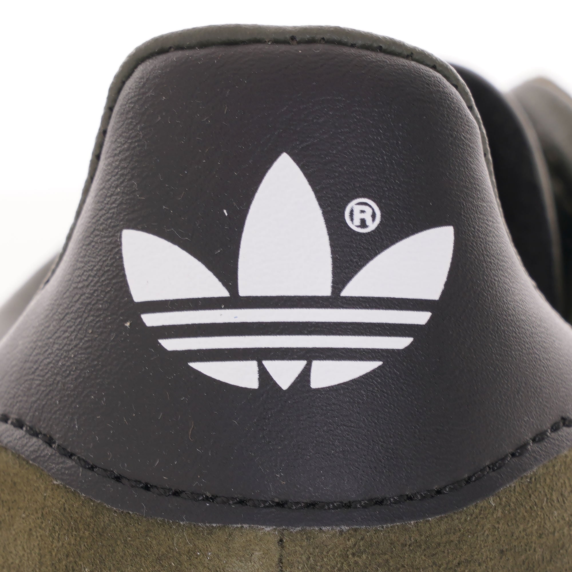 A close-up of the Adidas Trefoil logo on the black heel tab of an olive green trainer. 