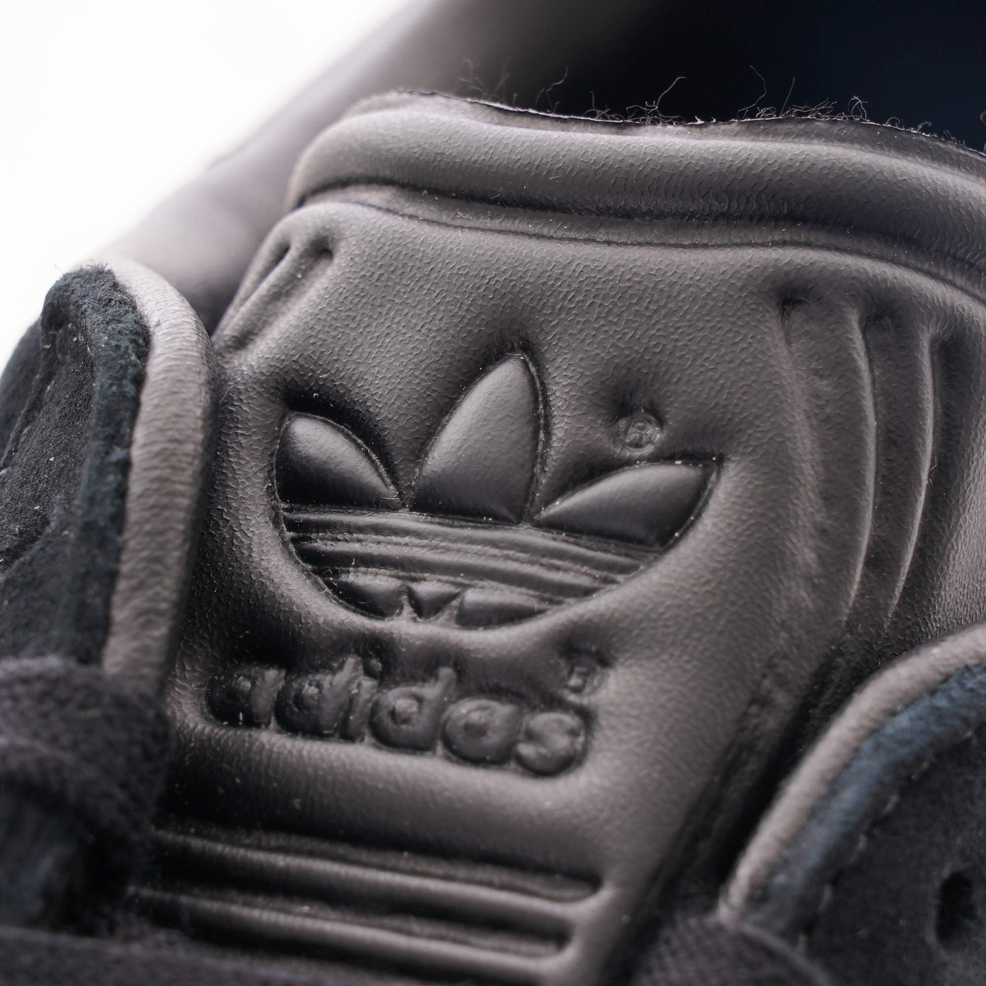 A close-up of the Adidas Trefoil logo embossed on the tongue of a black trainer.