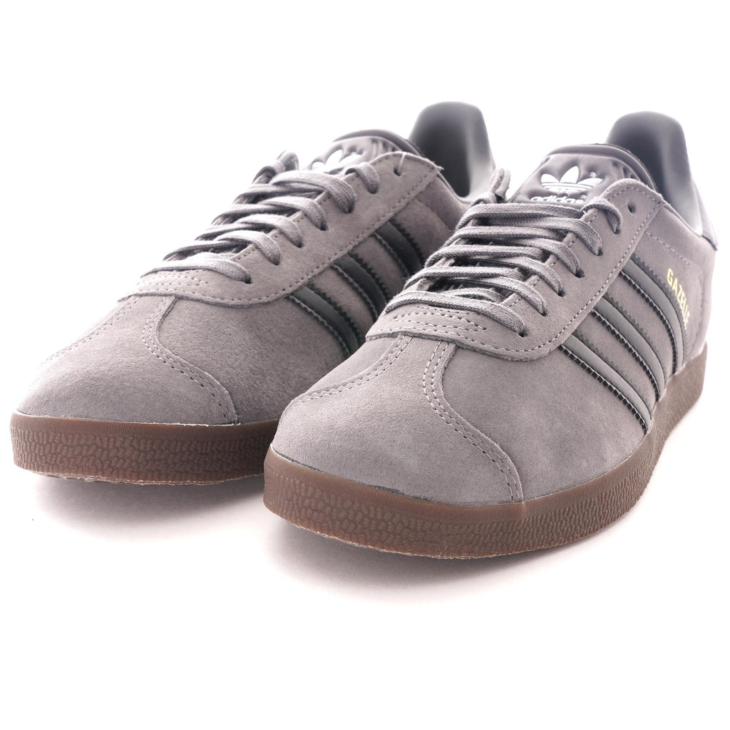 A pair of grey Adidas Gazelle trainers with black stripes and brown soles, viewed from an angle.