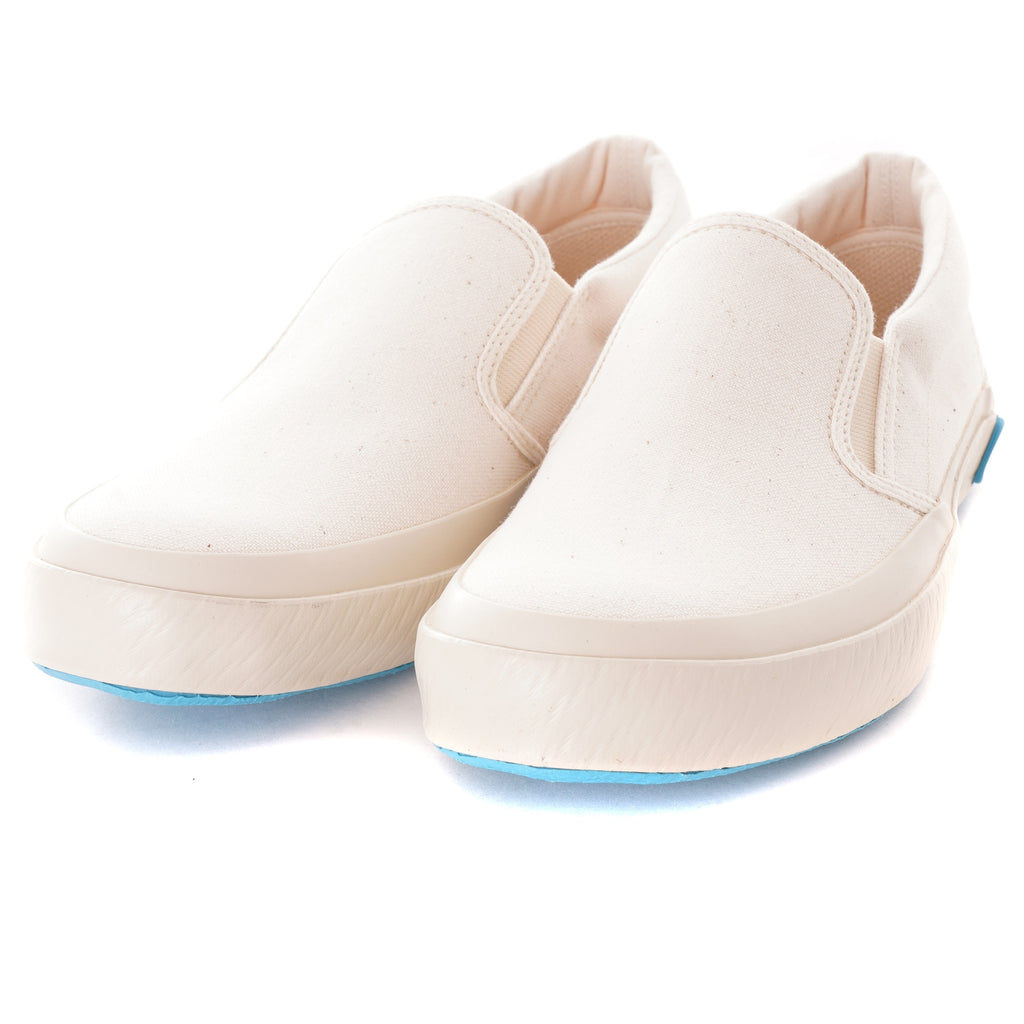 A pair of white slip-on canvas shoes with light blue soles are shown from an angle.