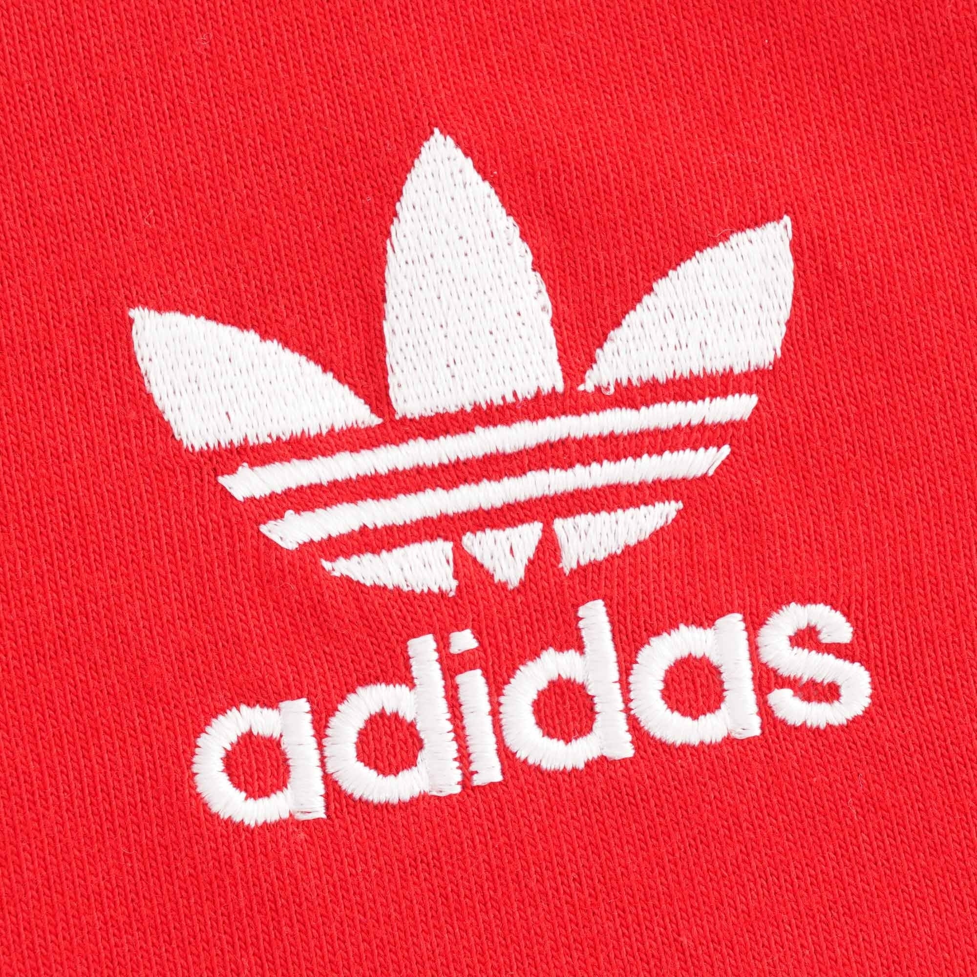 Close-up of the white embroidered Adidas Trefoil logo on the red tee.