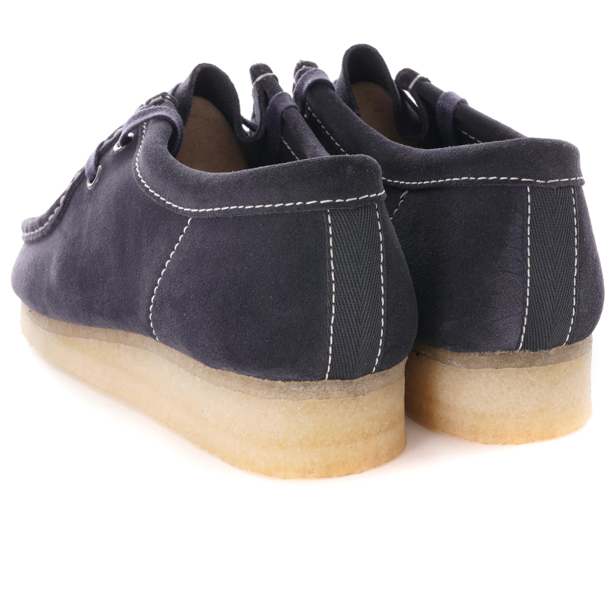 A pair of dark blue suede shoes with white stitching and light brown crepe soles, from behind.